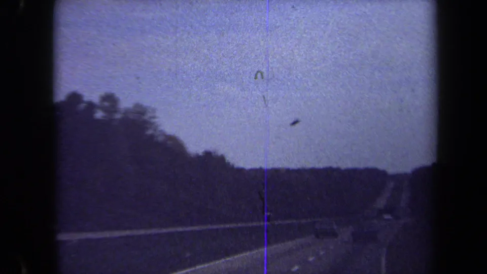 1970 8mm windshield view of a highway running through wooded terrain, with distant vehicles and visible film artifacts.