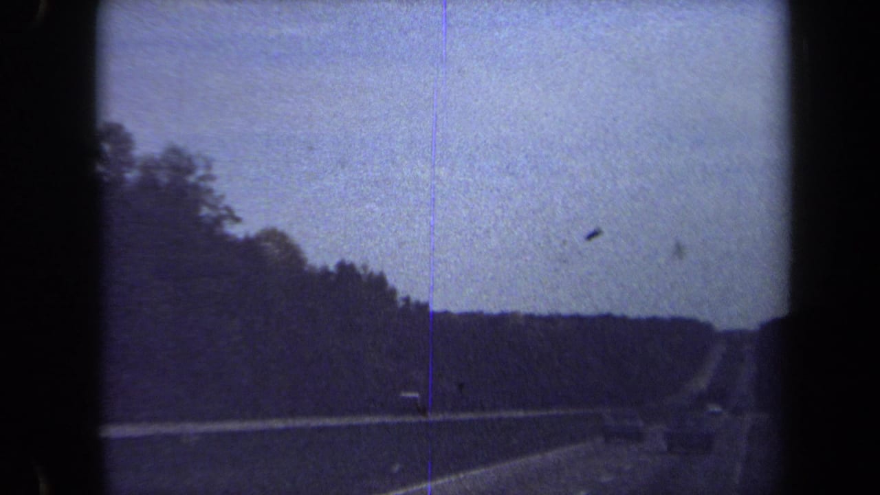 1970 8mm home-movie frame showing a dim passenger-side view of a highway bordered by trees near Washington, DC metadata location.