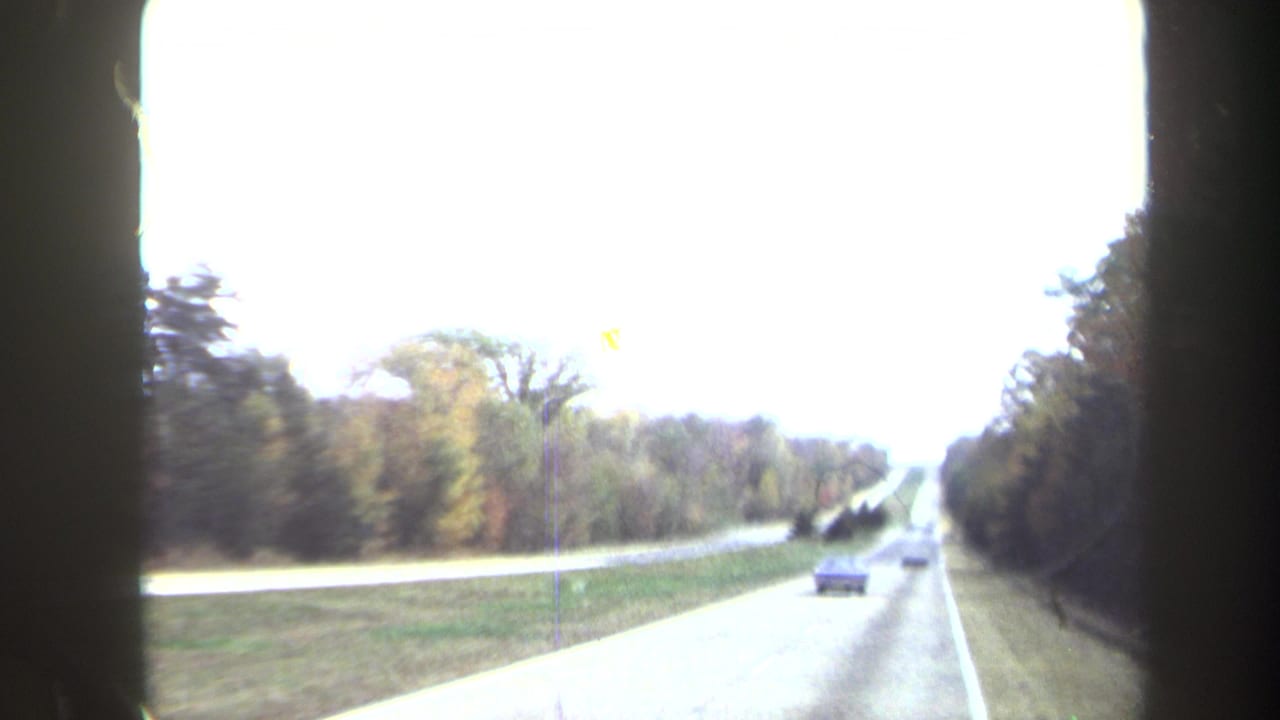 Bright 1970 8mm frame of a divided highway from inside a moving car, with vehicles ahead and trees along the roadway.