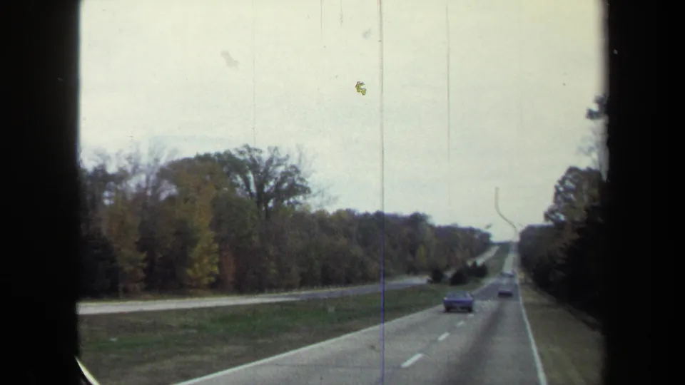 1970 8mm passenger-view frame showing a multi-lane highway, cars ahead, grassy median, and tree-lined roadside.