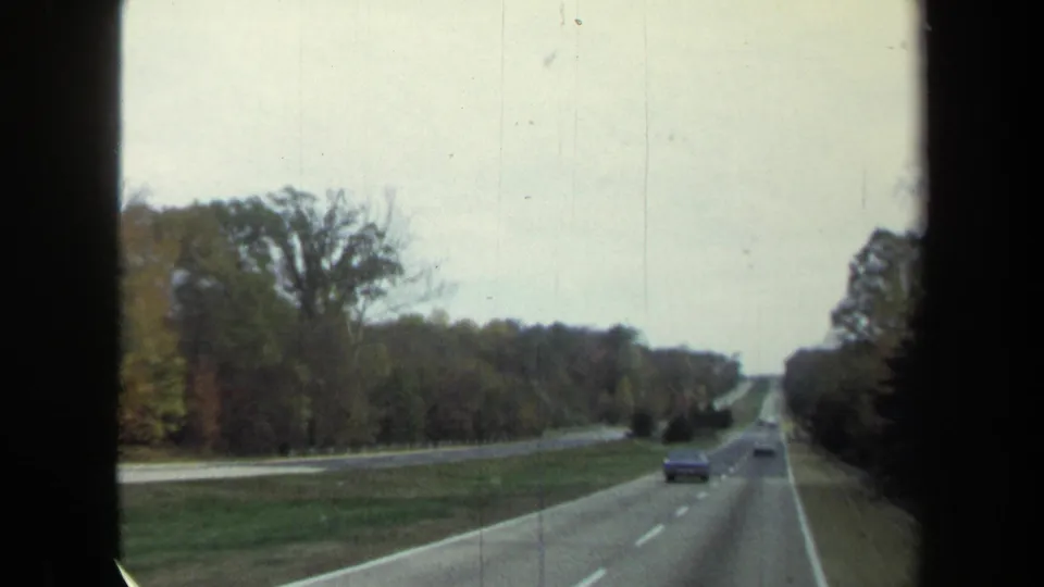 1970 8mm frame from a moving car showing a divided highway with vehicles ahead and wooded shoulders in the Washington, DC metadata area.