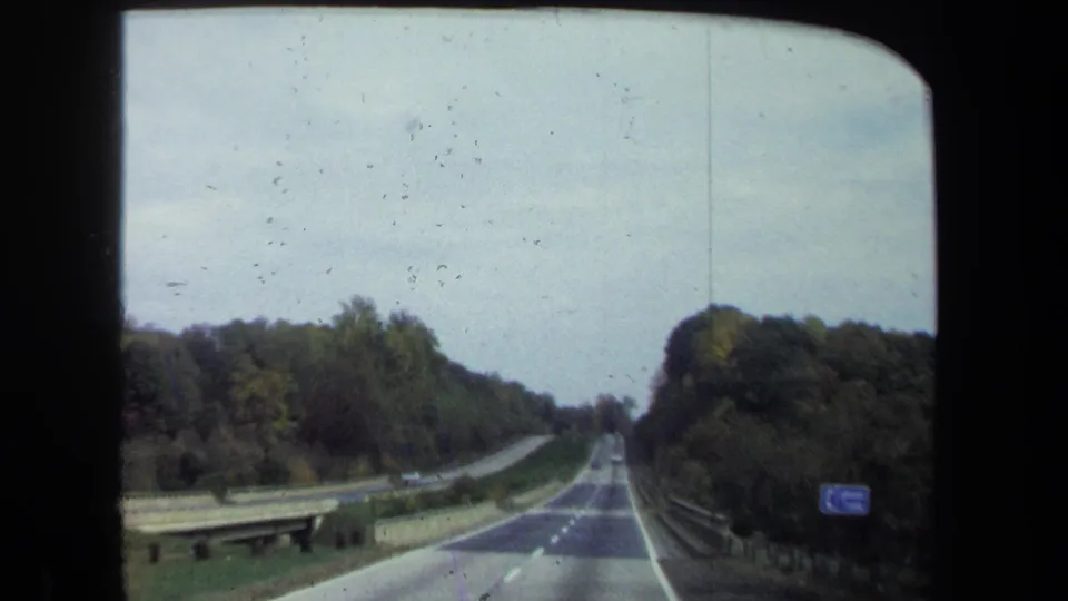 1970 8mm windshield view of a wooded two-lane road in the Washington, DC area, with guardrails, distant cars, and a blue roadside sign.