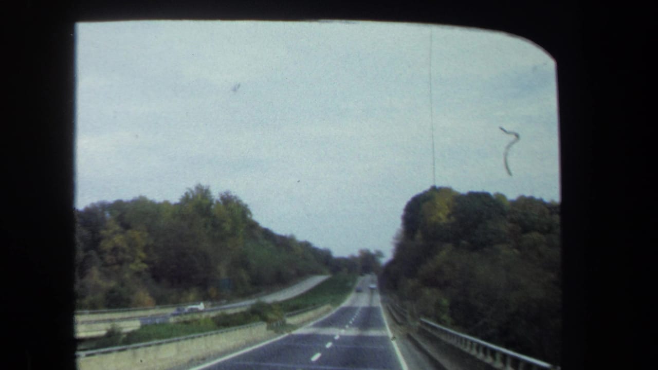 Windshield view on 8mm film of a two-lane highway through trees near Washington, DC, with guardrails and small cars ahead.