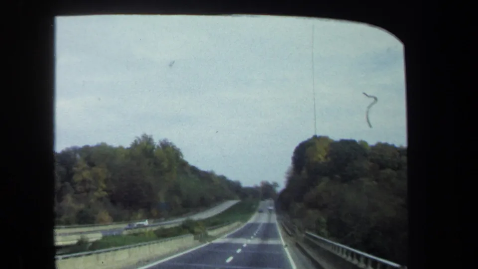 Windshield view on 8mm film of a two-lane highway through trees near Washington, DC, with guardrails and small cars ahead.