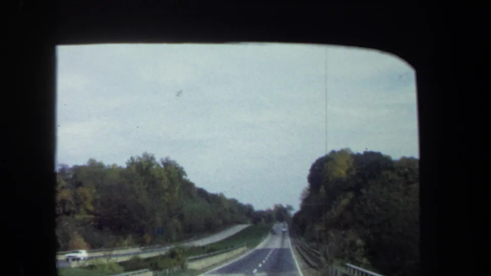 1970 8mm car-window view approaching an overpass or raised roadway through a wooded Washington, DC area road corridor.