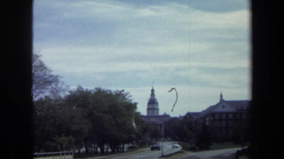 1970 Washington, DC roadway view with trees, passing cars, a distant dome, and a large civic building partly obscured by foliage.