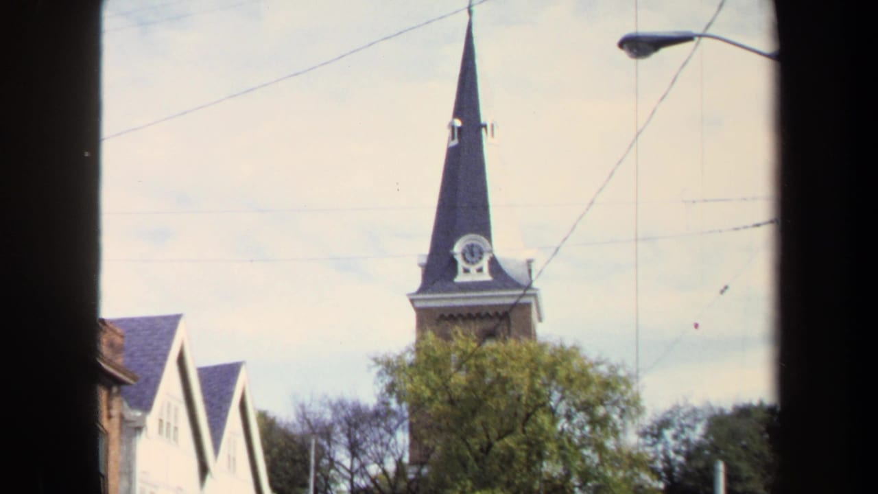 Washington, DC 1970 home-movie frame with a tall church steeple, clock face, trees, overhead wires, and a streetlight seen through a dark foreground edge.