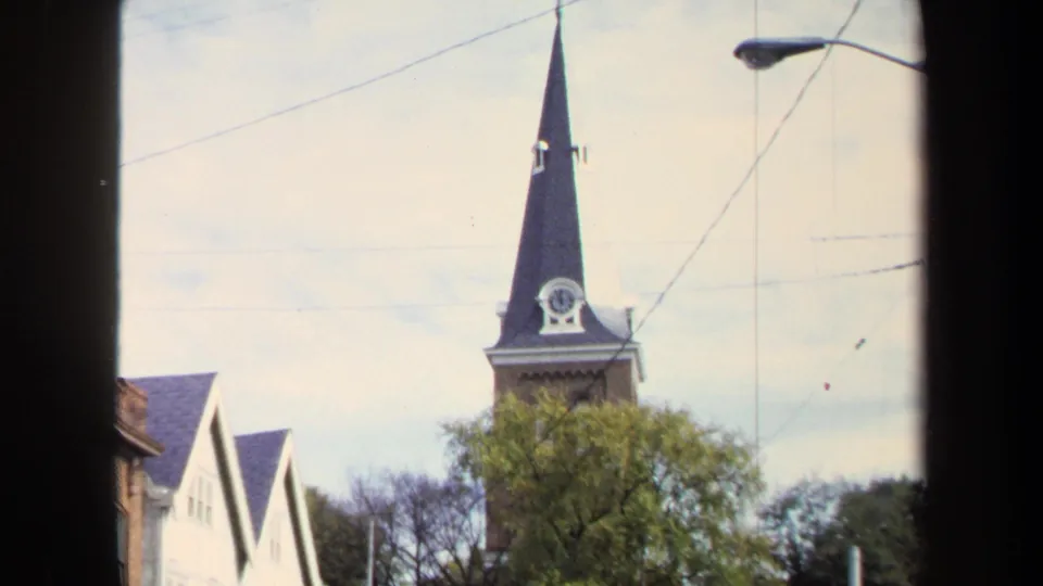 Washington, DC 1970 home-movie frame with a tall church steeple, clock face, trees, overhead wires, and a streetlight seen through a dark foreground edge.
