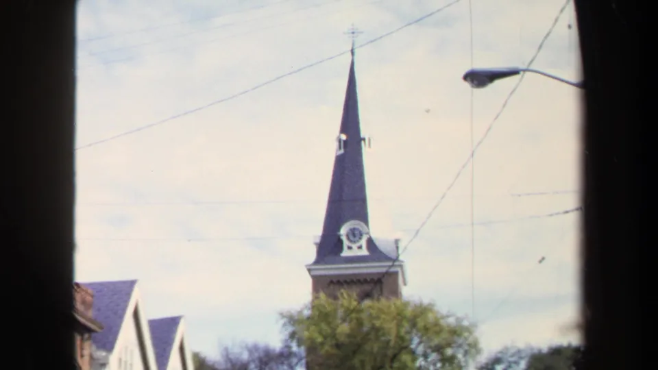 1970 Washington, DC frame showing the church steeple and clock above trees, with overhead wires, a streetlight, and the edge of a neighboring building.