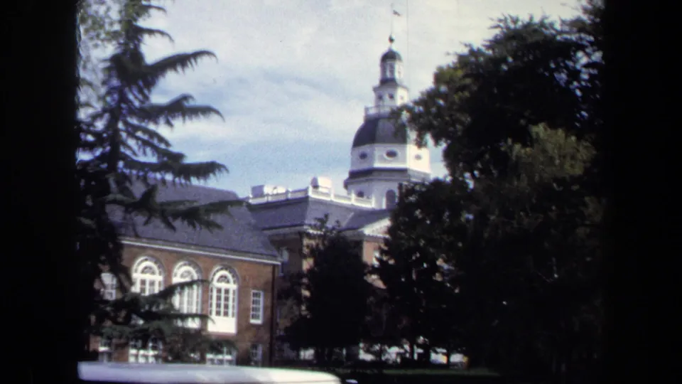 1970 Washington DC frame showing the domed tower rising above a brick building, with tree branches and a white car roof in the foreground.