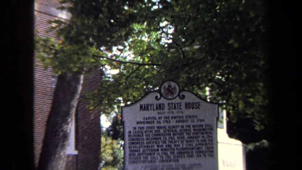 Close 1970 8mm view of the Maryland State House marker under leafy branches with brickwork behind it.