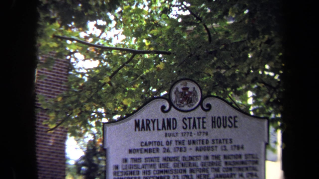 1970 8mm frame of the Maryland State House historical marker seen through leafy trees, with brickwork behind it.