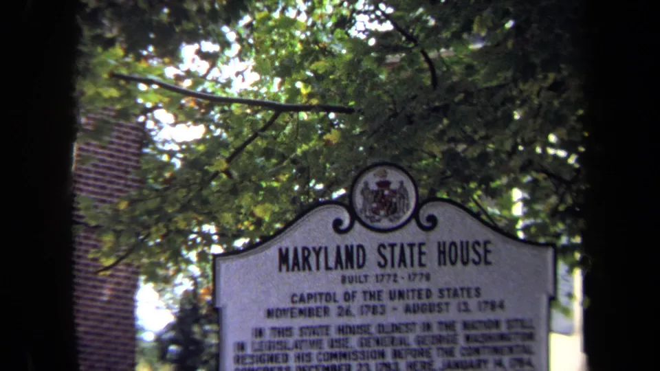 1970 8mm frame of the Maryland State House historical marker seen through leafy trees, with brickwork behind it.