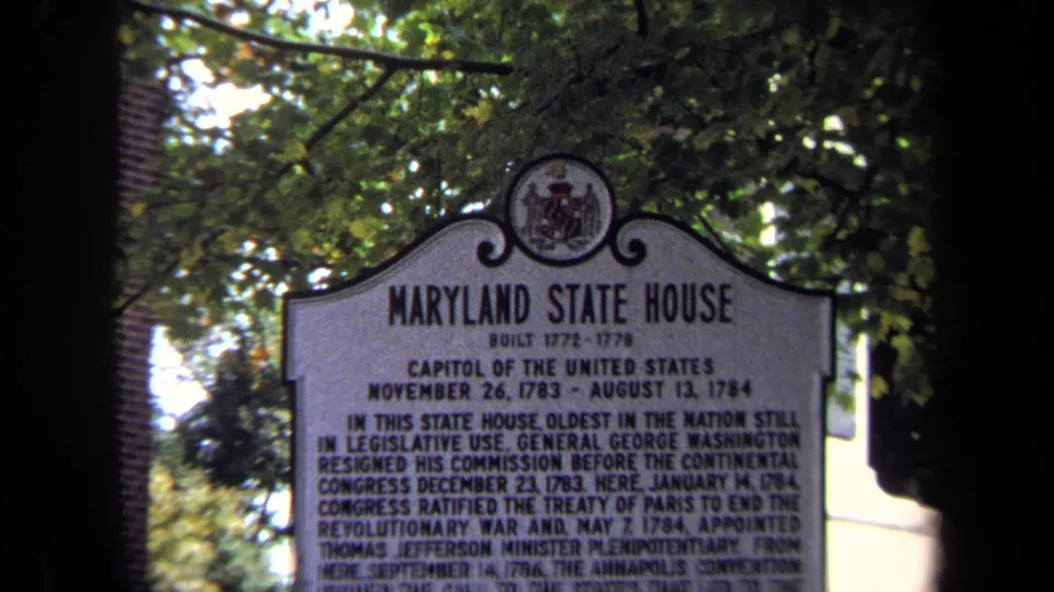 1970 8mm frame with the Maryland State House marker filling most of the view, surrounded by tree leaves and brick architecture.