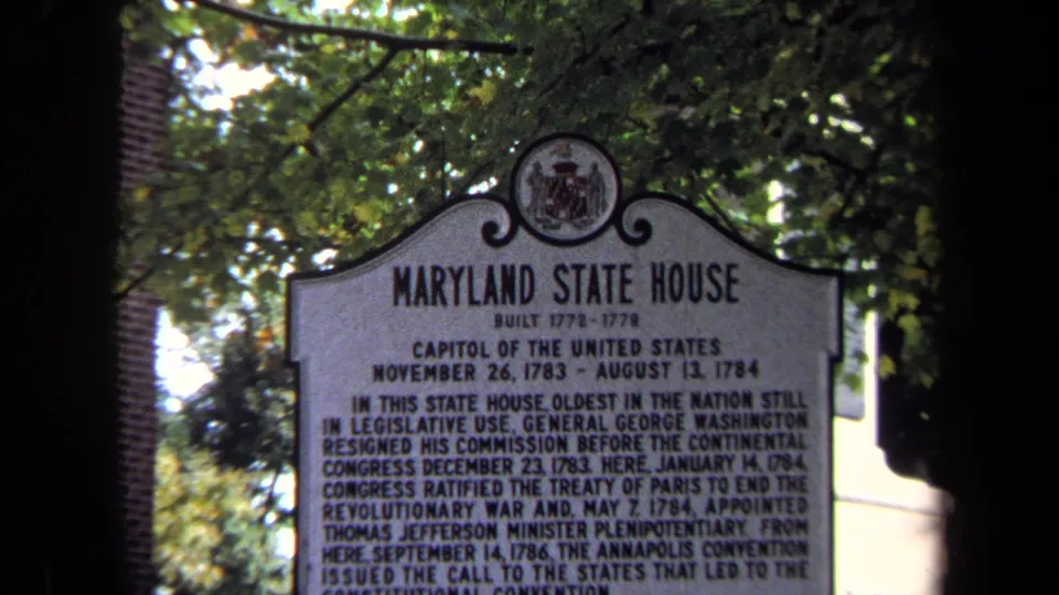 1970 8mm frame of a Maryland State House historical marker seen through foliage and shadowed foreground framing.