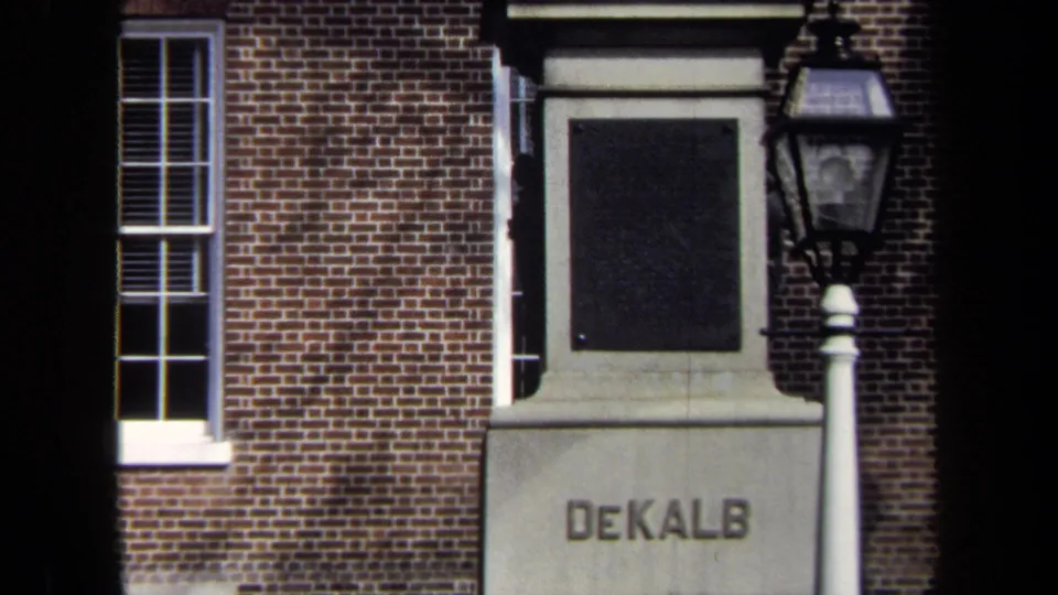 1970 8mm view of a brick civic building facade with a window, lamp post, and DeKalb monument pedestal.