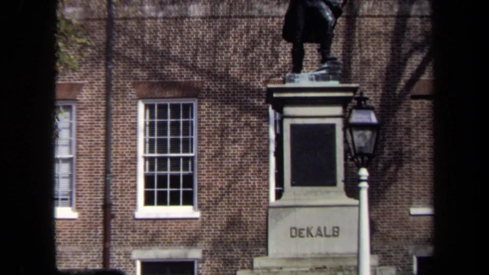 1970 8mm frame showing the DeKalb monument area against a brick facade, with a statue base and lamp post visible.