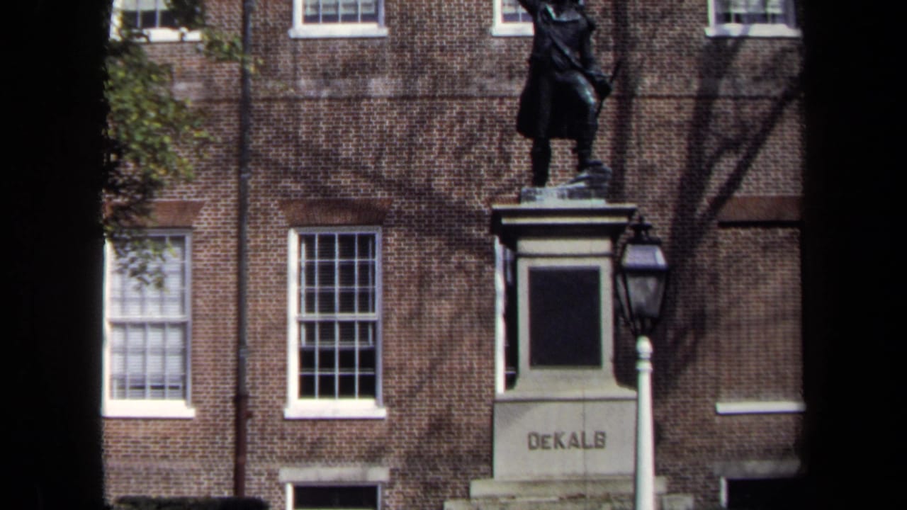 1970 Washington, DC 8mm frame showing a DeKalb pedestal statue before a brick facade with sash windows and tree shadows.