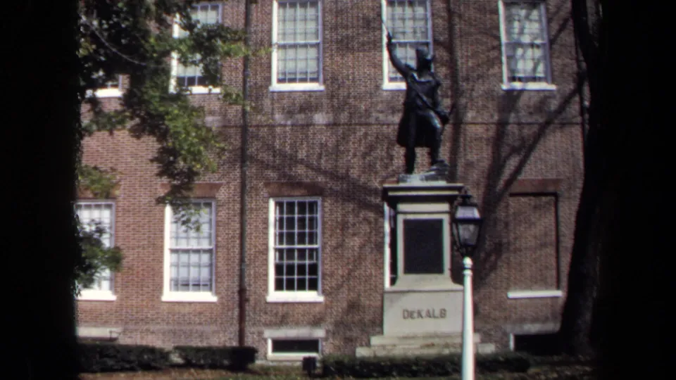 1970 Washington, DC home-movie frame with the DeKalb statue, a lamppost, brick facade, windows, and tree shadows.
