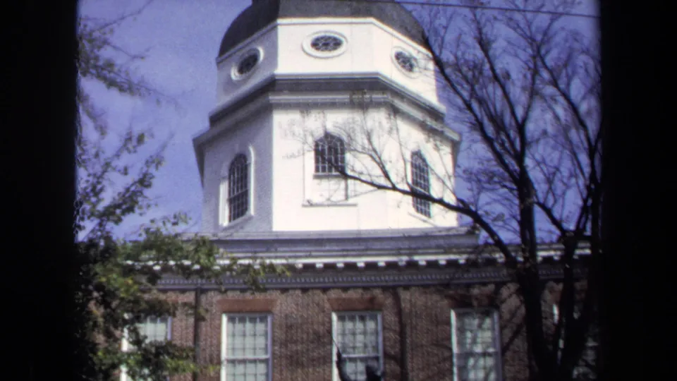 1970 Washington DC 8mm view looking up at a white church tower with arched windows, brick lower wall, branches, and pale blue sky.
