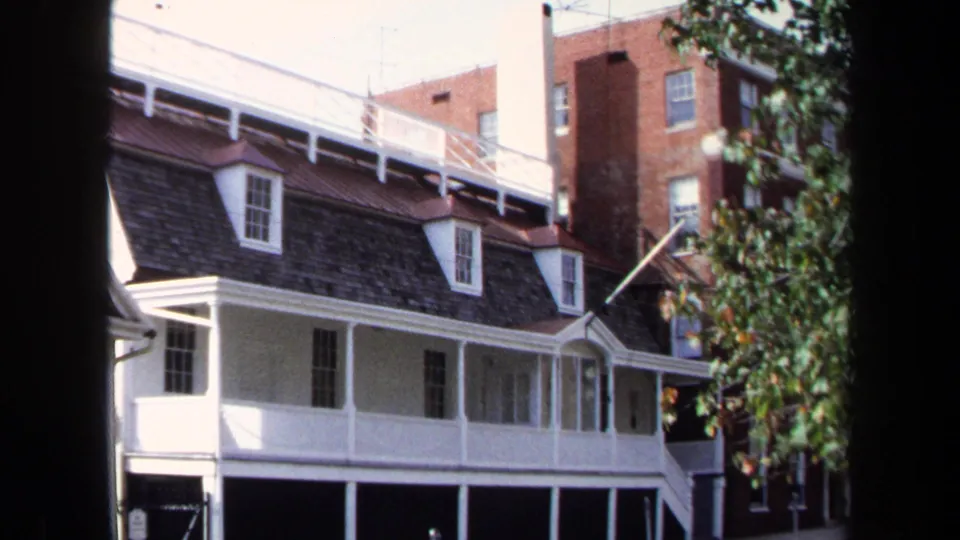 1970 Washington, DC frame showing the Shaw House exterior with white porch columns, dark shingled roof, trees, and urban buildings nearby.