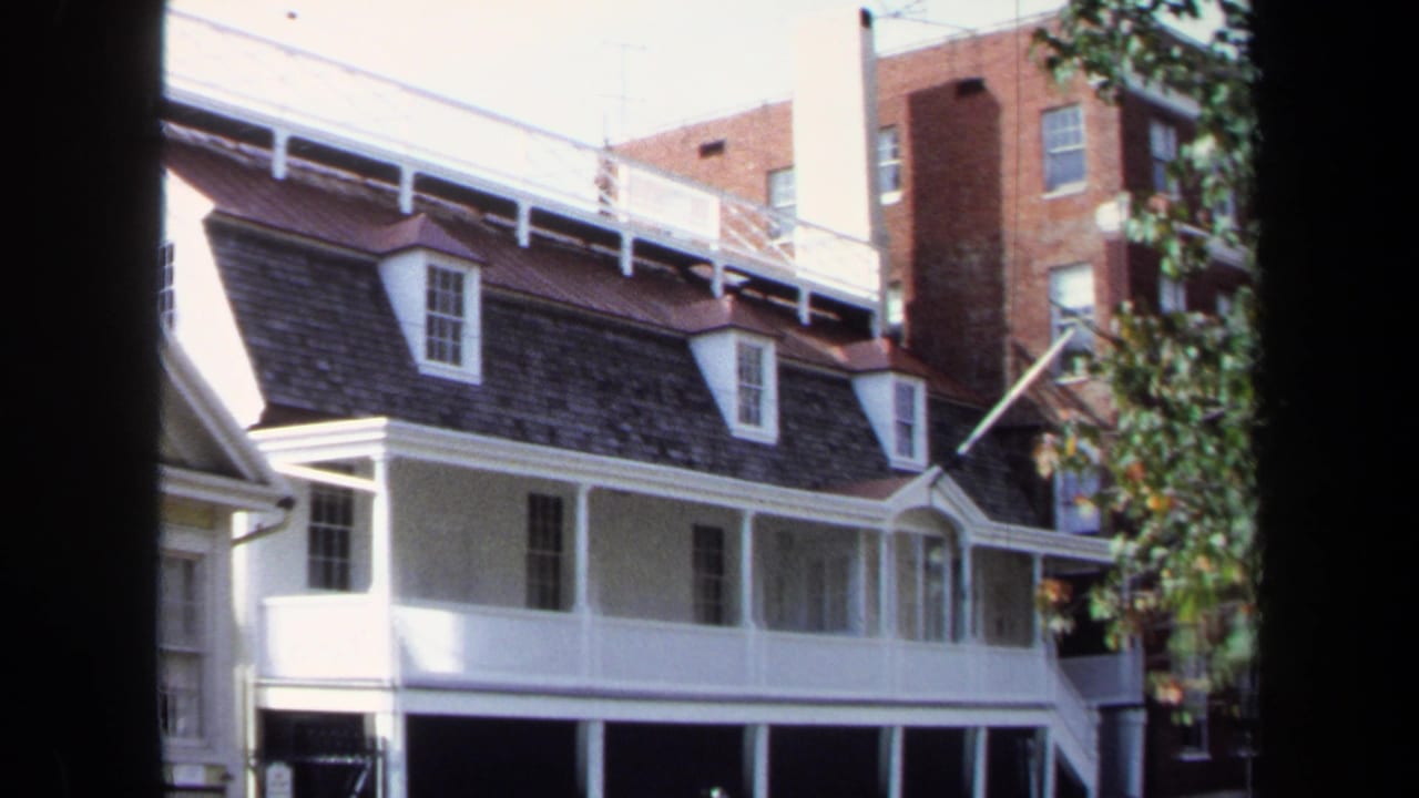 1970 Washington, DC home-movie frame showing a white residential facade with dormer windows, porch supports, and a brick building in back.