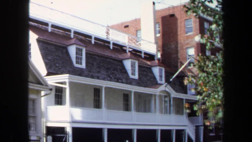 1970 Washington, DC home-movie frame showing a white residential facade with dormer windows, porch supports, and a brick building in back.