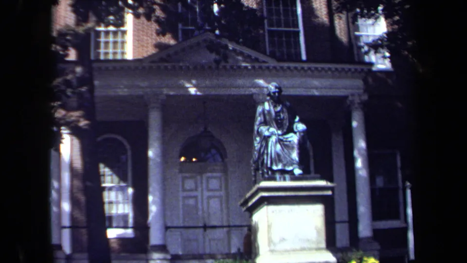 Seated statue and stone base seen in front of a classical Washington, DC building, partly shaded by trees.