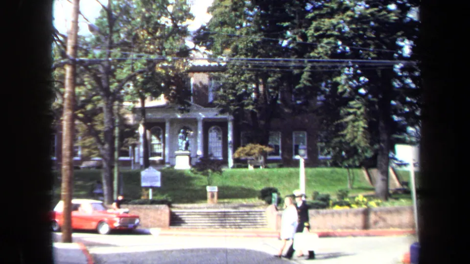 Wide 1970 Washington, DC street view with the same monument across a lawn, pedestrians on the sidewalk, and a red car at left.