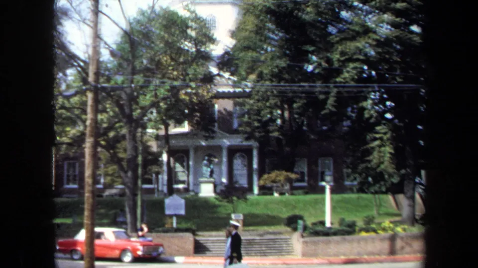 1970 Washington, DC home-movie frame showing pedestrians, a red car, trees, steps, and the monument set back from the street.