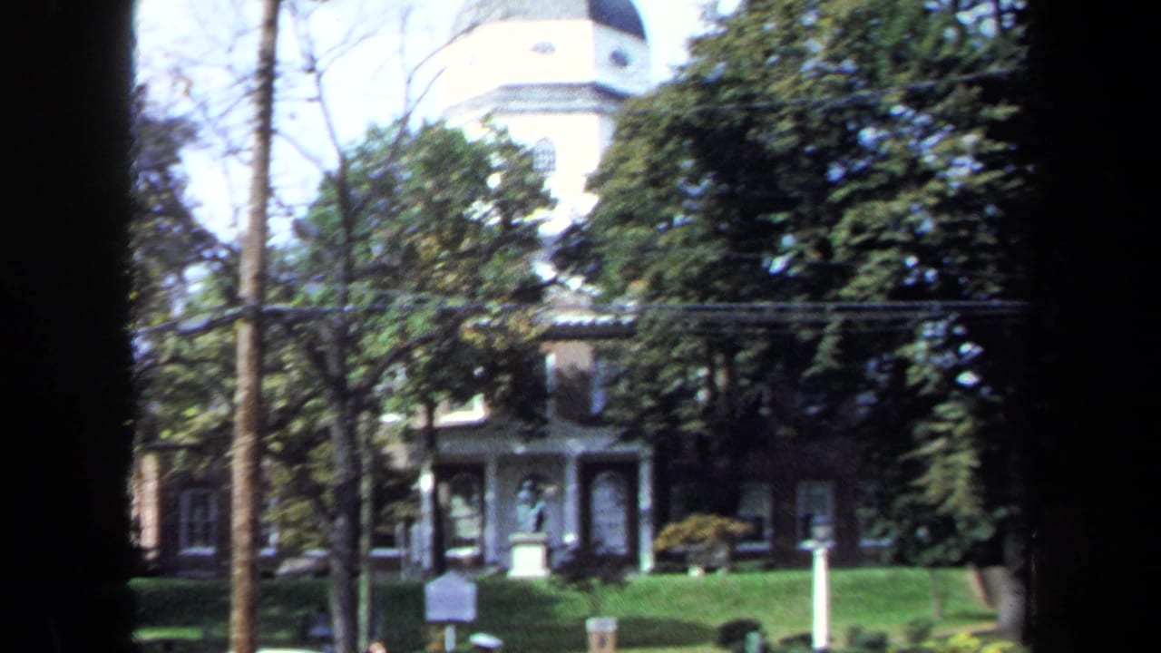 1970 8mm view of a historic brick exterior with a white cupola seen through trees and handheld film edges.