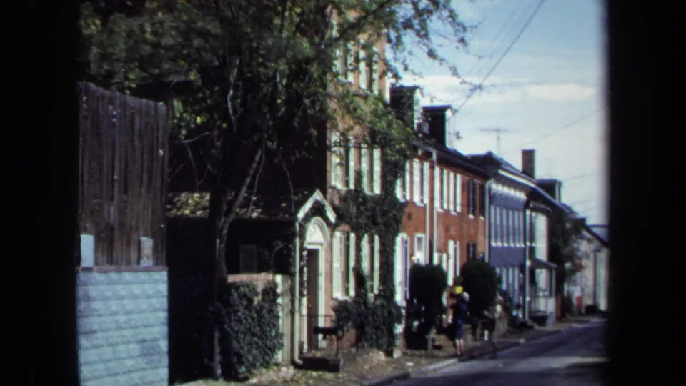1970 Washington, DC street view with brick row houses, trees, overhead wires, sidewalk, and distant pedestrians.