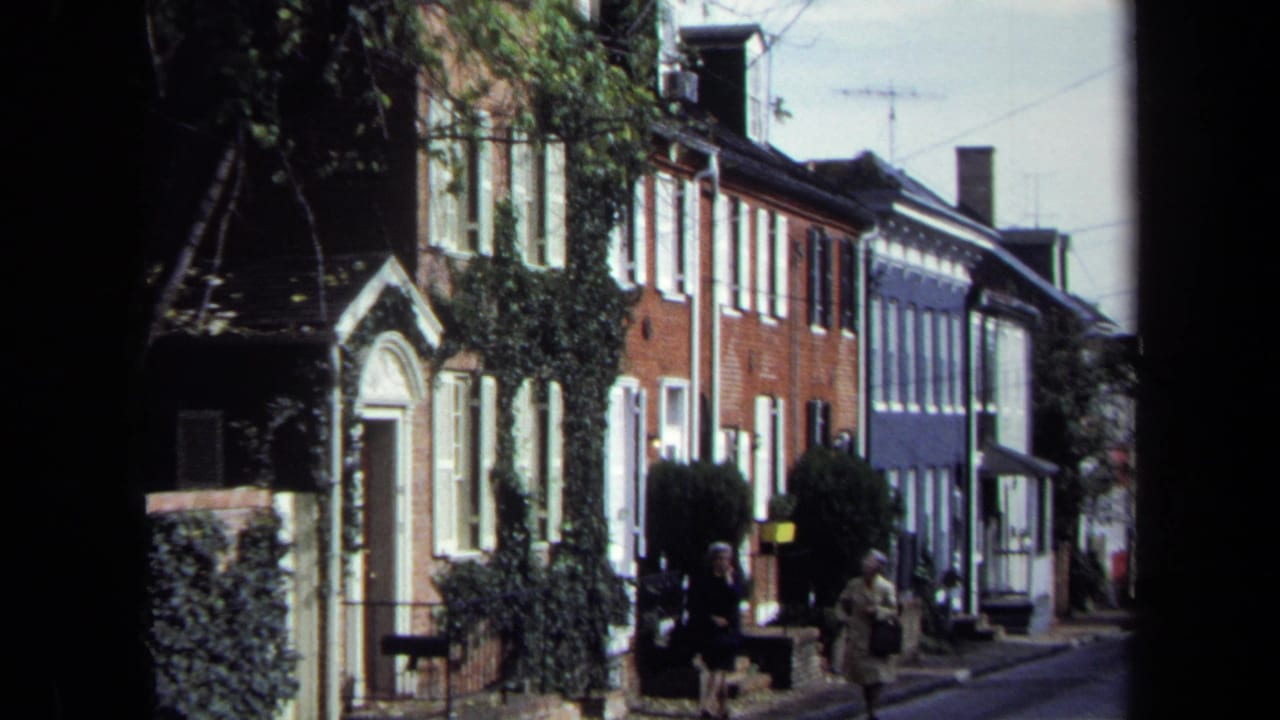 1970 Washington, DC street view with two women walking past brick and painted row houses in sunlight.
