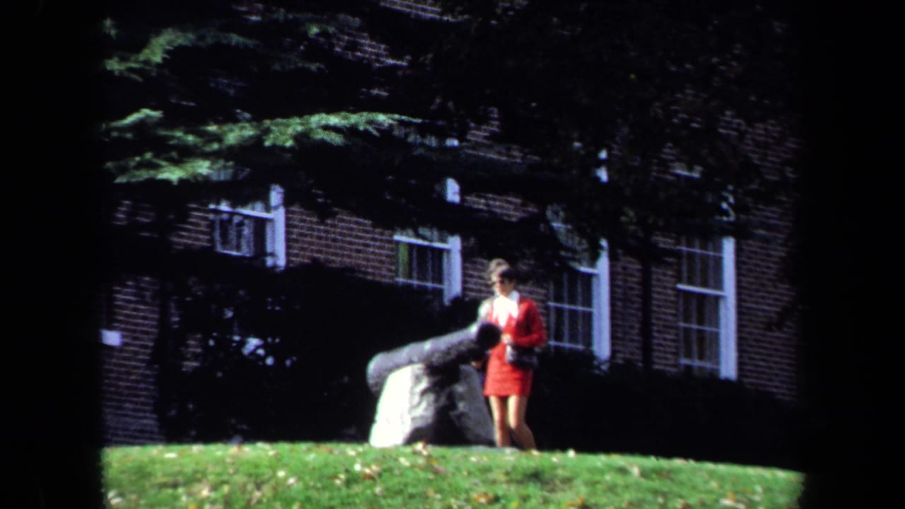 1970 Washington, DC campus scene with a student in red near a large cannon, brick building, windows, and trees behind her.