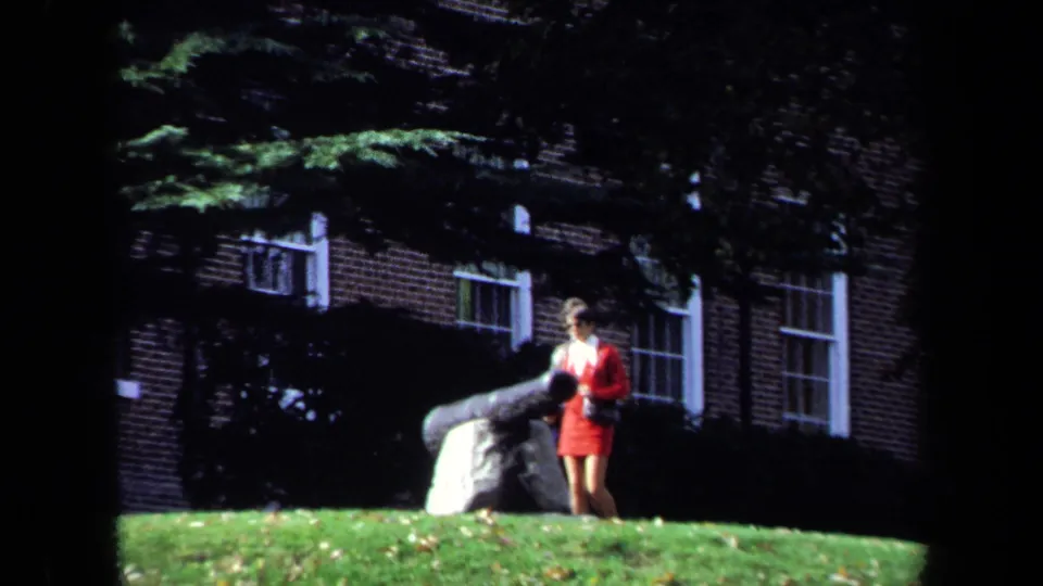 1970 Washington, DC campus scene with a student in red near a large cannon, brick building, windows, and trees behind her.