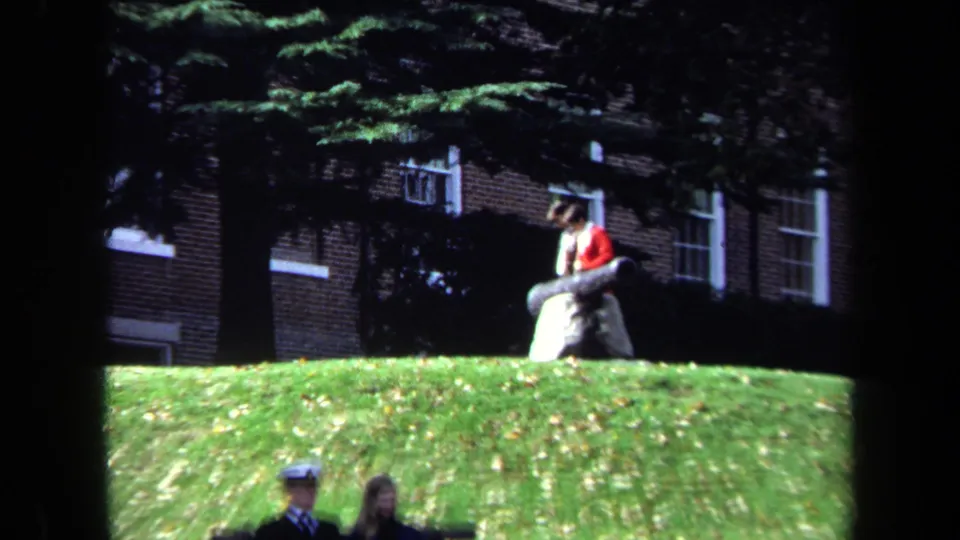 Campus footage shows a student near the cannon on the hill while two people walk through the lower foreground in partial view.