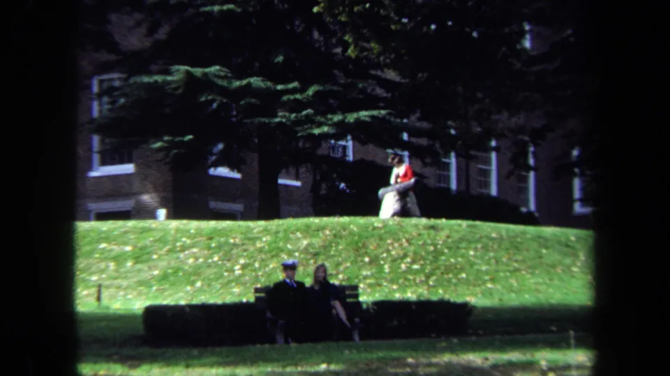 Two people cross the foreground near a hedge while the cannon and a seated student remain visible on the campus slope above.