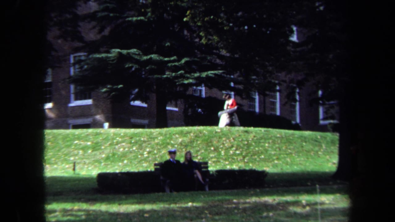 1970 Washington, DC home-movie frame showing people seated on benches across a green lawn with trees and brick buildings behind them.