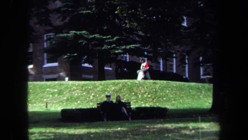 1970 Washington, DC home-movie frame showing people seated on benches across a green lawn with trees and brick buildings behind them.
