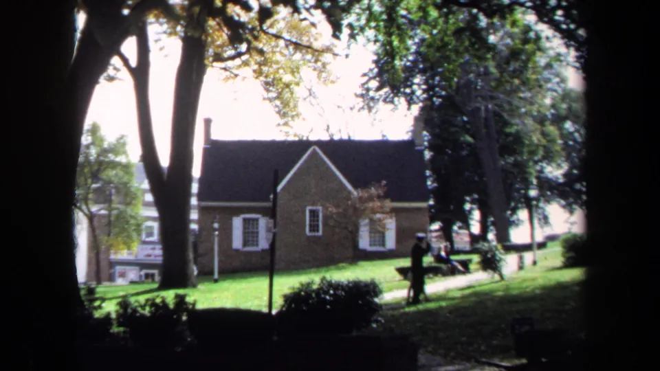 Wide 1970 Washington, DC park frame with a brick building, tall trees, a bench, and a person walking across the lawn area.