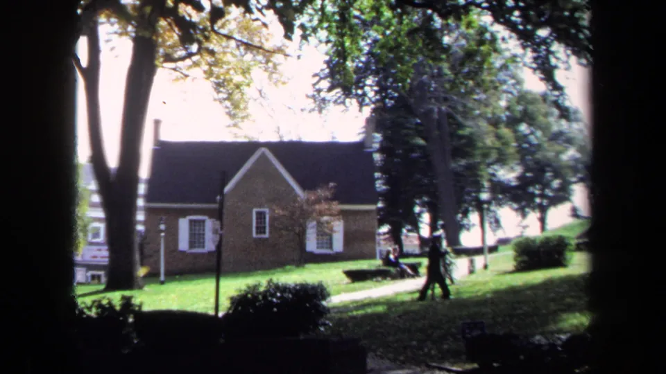 The seated couple remains near the path as another person walks through the 1970 Washington, DC park scene with trees and brick buildings.