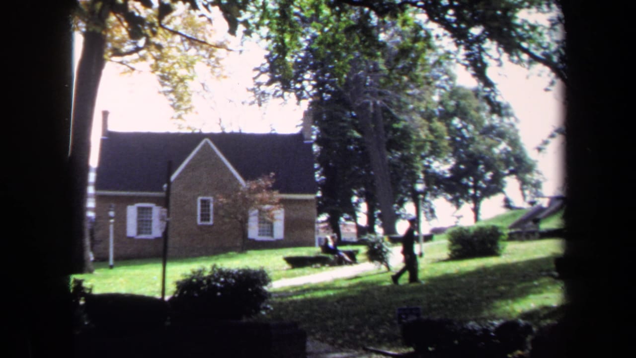 1970 Washington, DC home-movie frame showing a brick building, white shutters, lawn, benches, and trees.