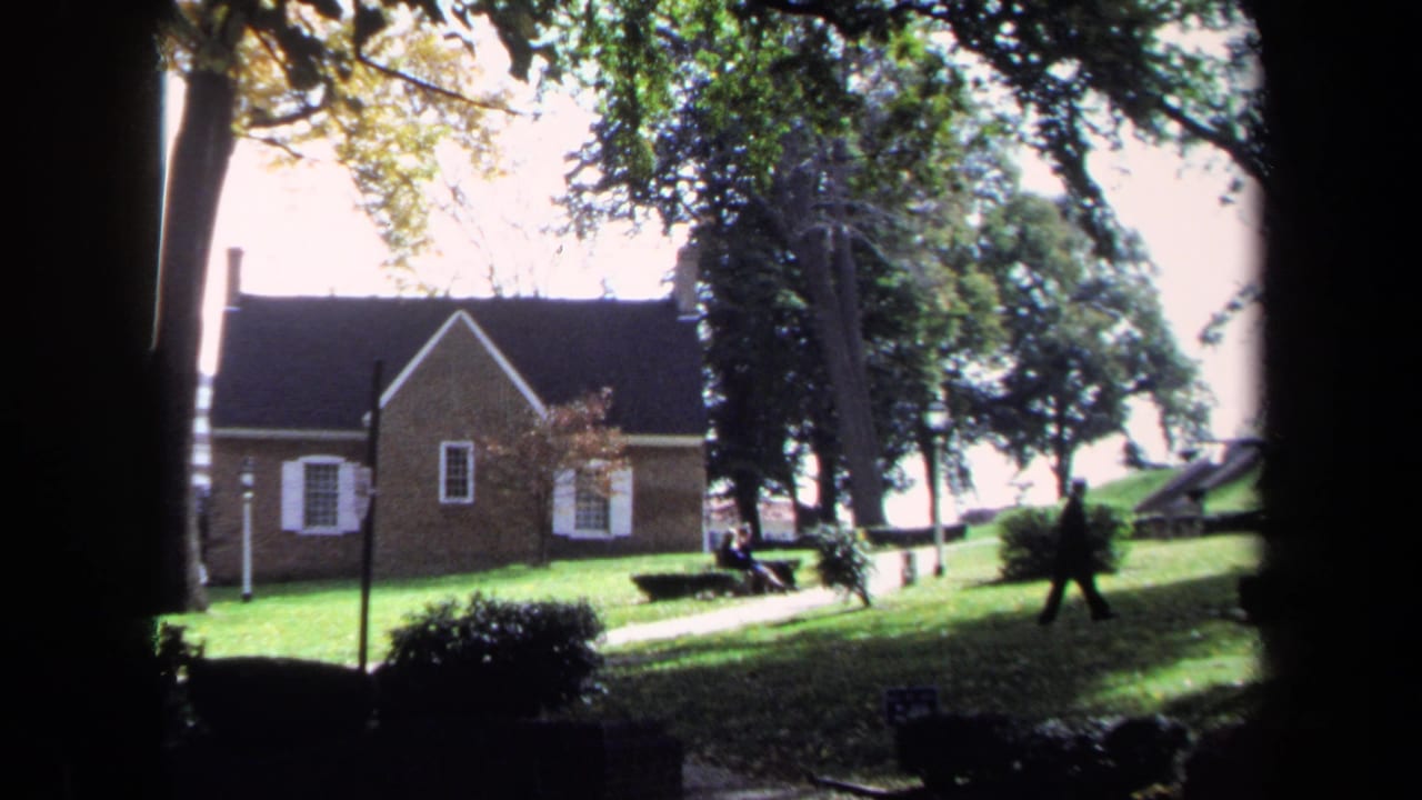 Person walking across a grassy area near a brick building and trees.