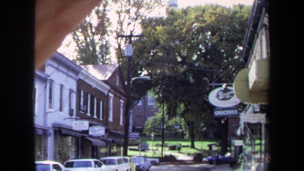 1970 Washington, DC home-movie frame showing small storefronts, parked cars, leafy trees, and a visible groceries sign from a shaded window view.