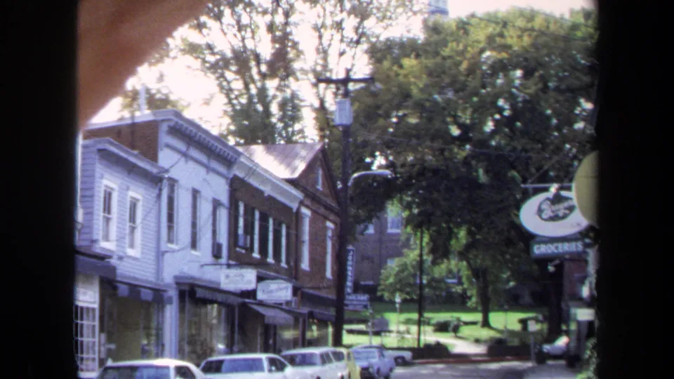 1970 Washington, DC street scene seen from indoors, with low-rise shops, parked cars, trees, and a round groceries sign near the right side.