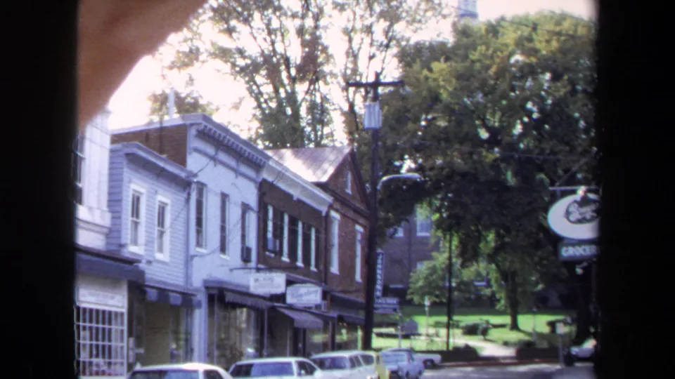 1970 Washington, DC home-movie view of a neighborhood block with storefront awnings, older cars, trees, and partially visible shop signage.