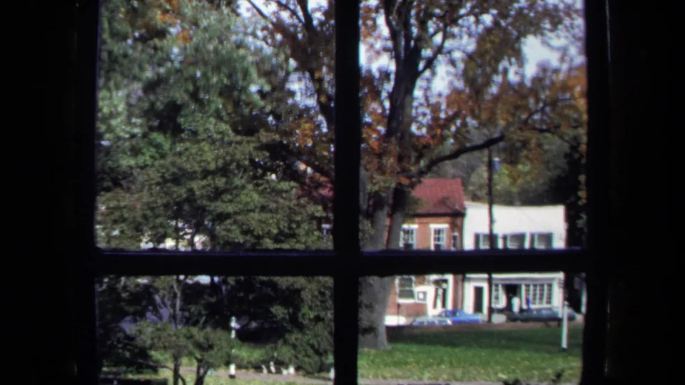 1970 Washington, DC home-movie frame looking through dark window muntins toward trees, grass, a small building, and a parked car.