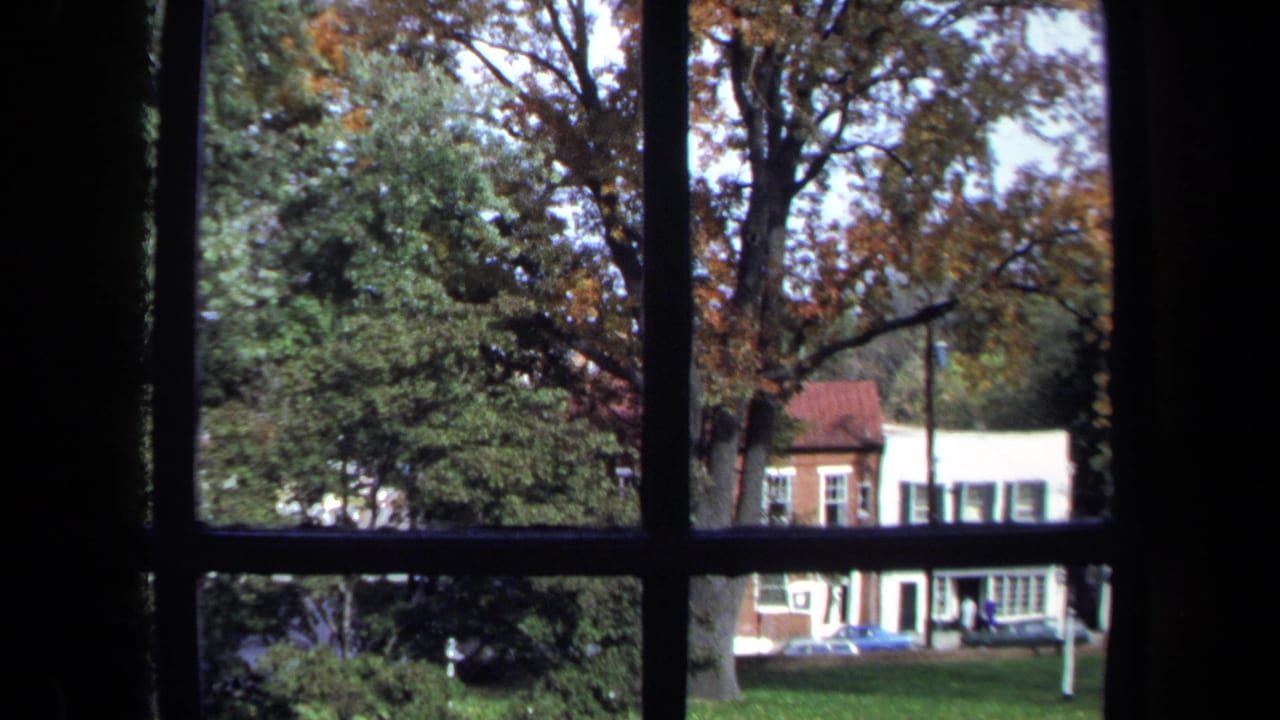 1970 Washington, DC home-movie frame looking through a dark-paned window at autumn trees and buildings across a lawn.