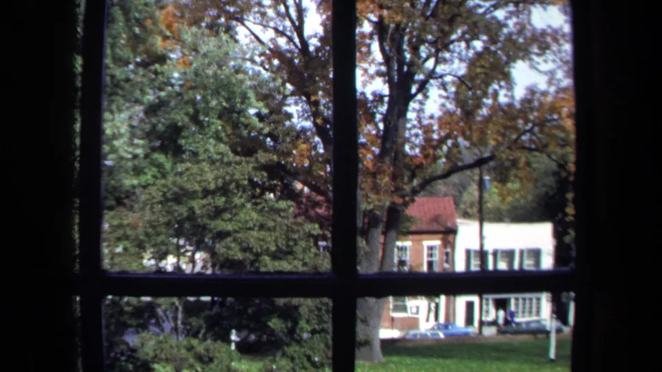 1970 Washington, DC home-movie frame looking through a dark-paned window at autumn trees and buildings across a lawn.