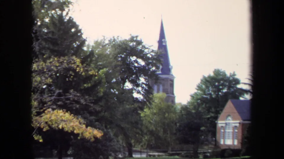 1970 8mm Washington, DC campus exterior with trees, autumn leaves, a steeple, and a brick building seen from indoors.